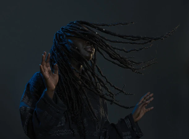 Black woman with dreadlocks flowing across her face wearing a dark patterned shirt against a dark background, photographed by Ian & Erick.