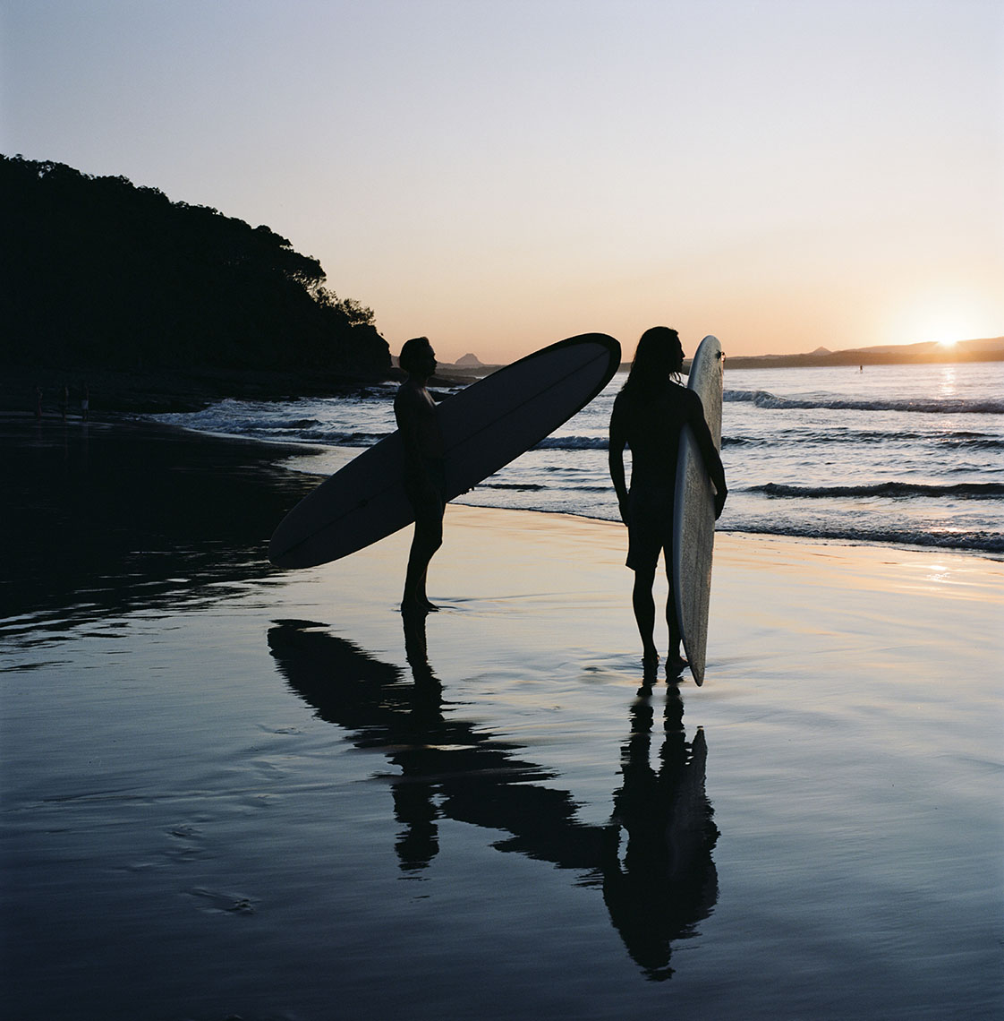 Two silhouetted surfers standing on a beach at sunset holding surfboards, with reflections in the wet sand.