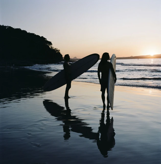 Two silhouetted surfers standing on a beach at sunset holding surfboards, with reflections in the wet sand.