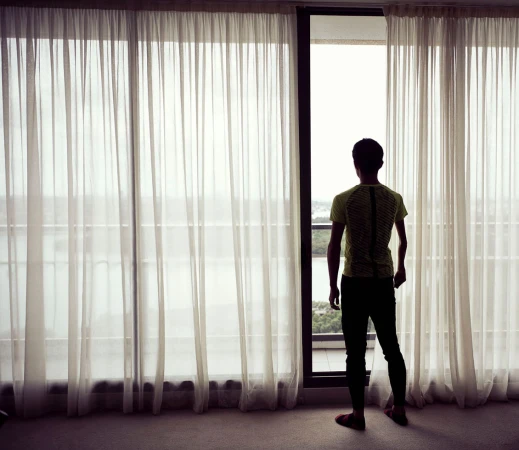 Rear view of a person looking out a window with sheer white curtains in a room with soft light.