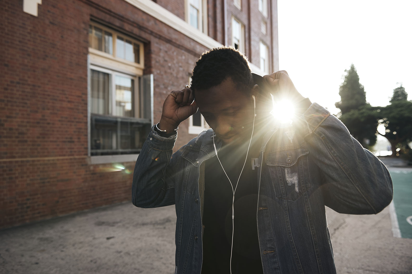 Black man wearing a denim jacket adjusting his hood against a brick building exterior in Sydney