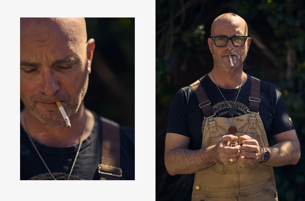 Portrait of a bald, middle-aged man smoking a cigarette, wearing glasses and brown overalls in a natural outdoor setting with dark foliage.