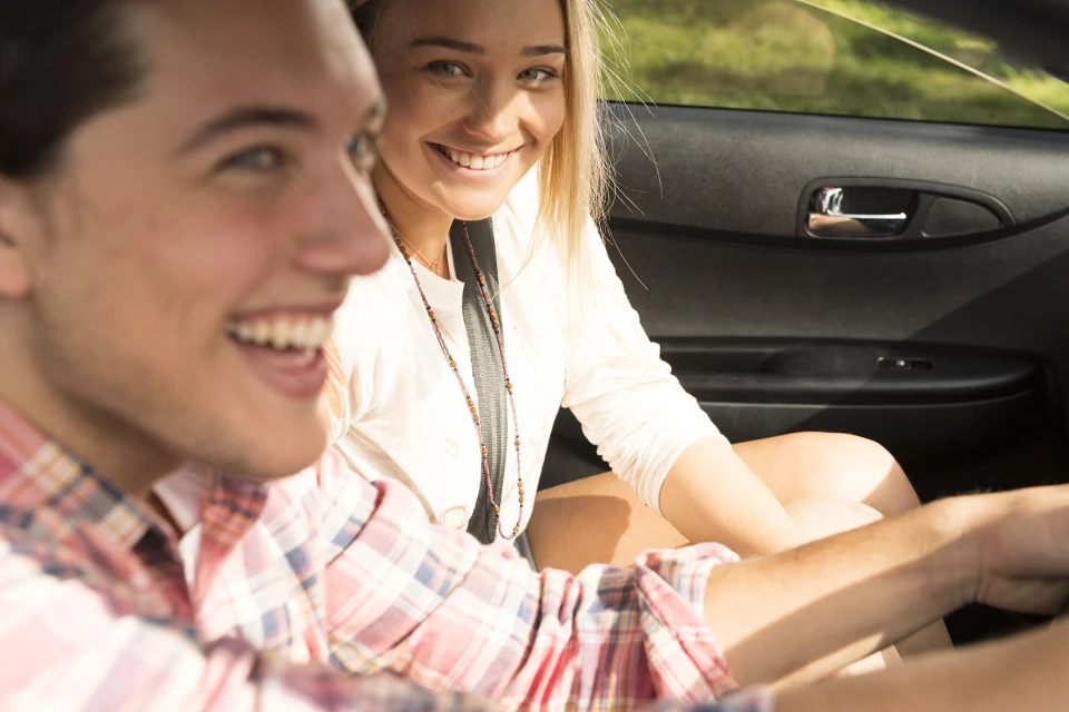 Smiling young couple in a car: a blond woman in the passenger seat and a brown-haired man driving with a pink and white plaid shirt.