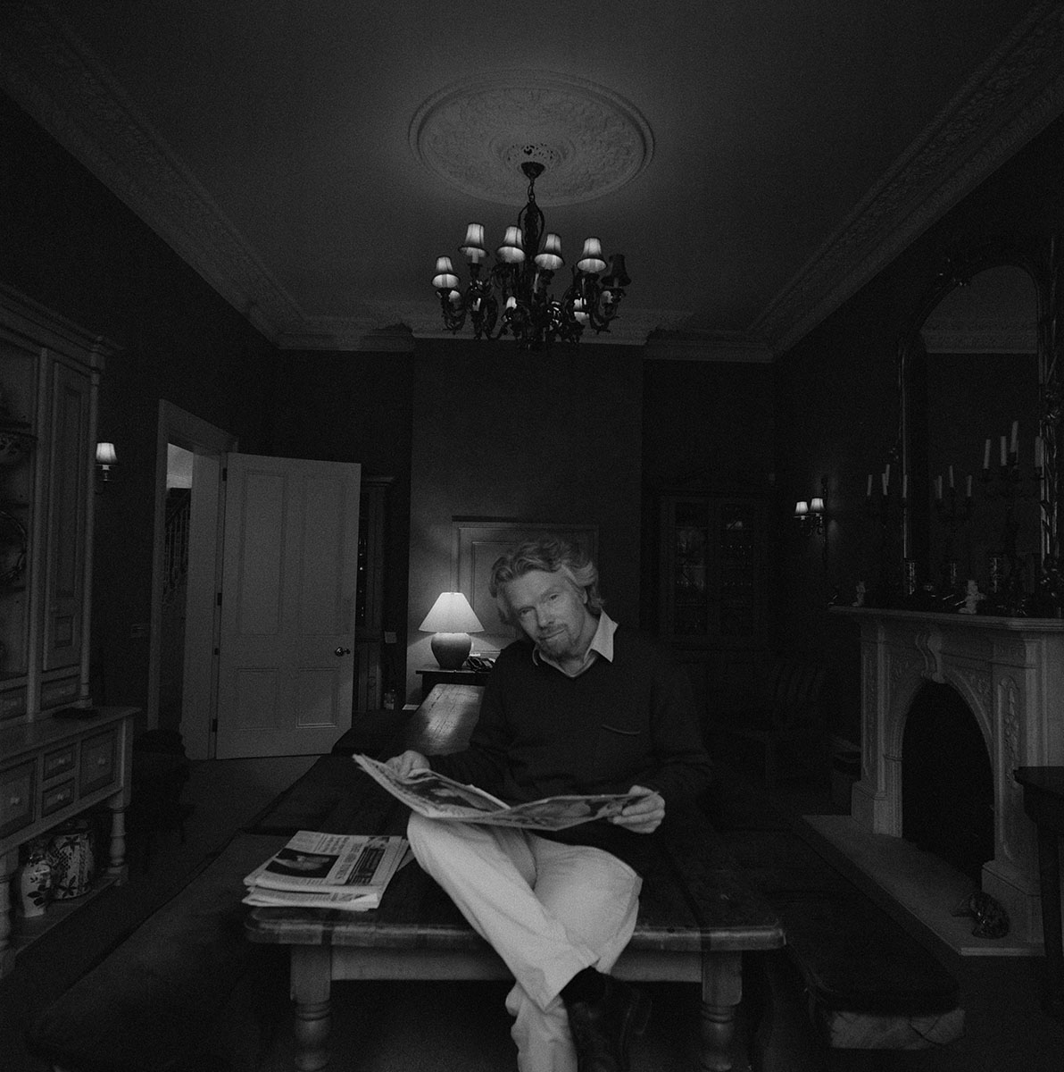 Black-and-white shot of a man with a newspaper sitting on a wooden table in a dark, ornately decorated room with a chandelier.