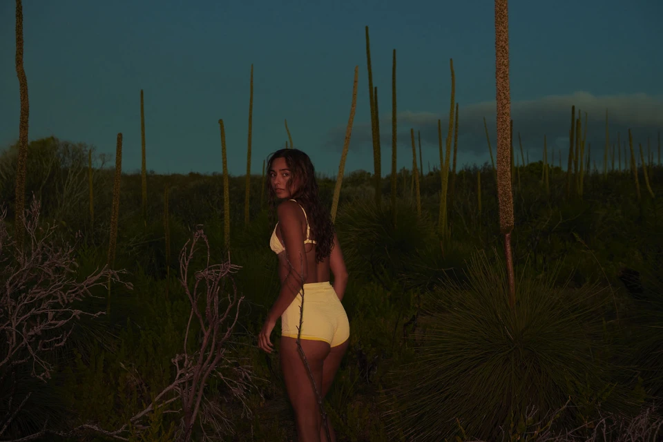 A young woman with long brown hair is seen in a yellow swimsuit with a light blue sky and tall reeds in the background.