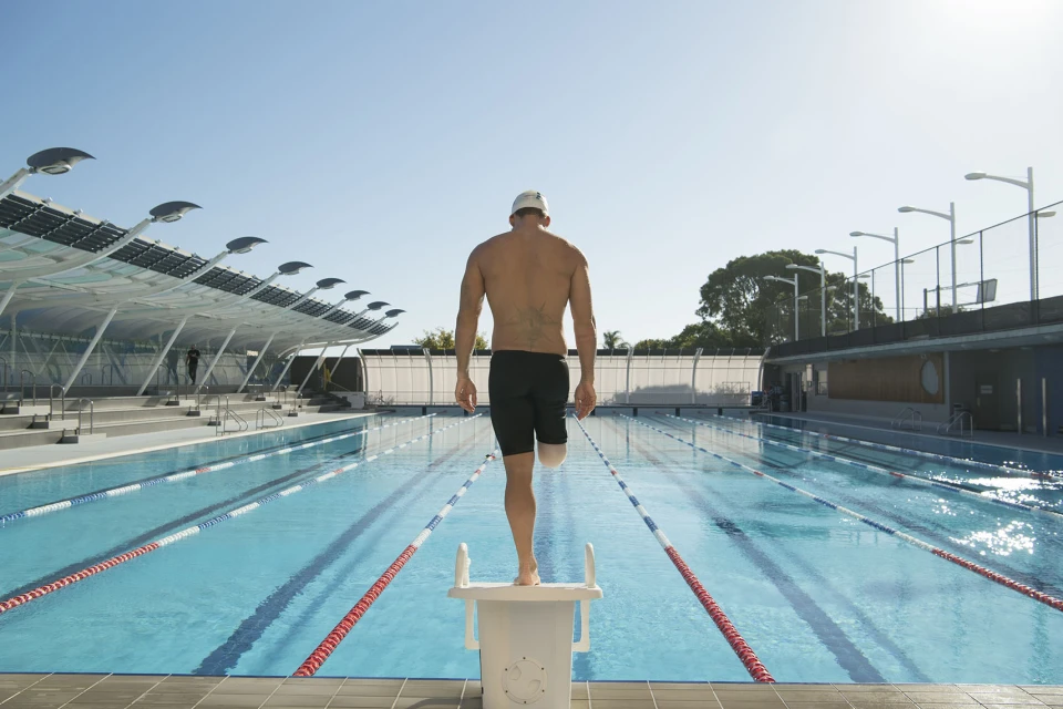 Adaptive swimmer stands on starting block with prosthetic leg, back view, outdoors on sunny day at outdoor swimming pool.