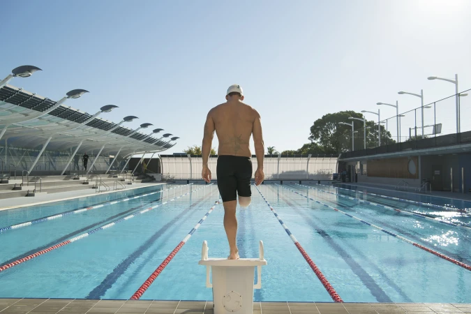 Adaptive swimmer stands on starting block with prosthetic leg, back view, outdoors on sunny day at outdoor swimming pool.