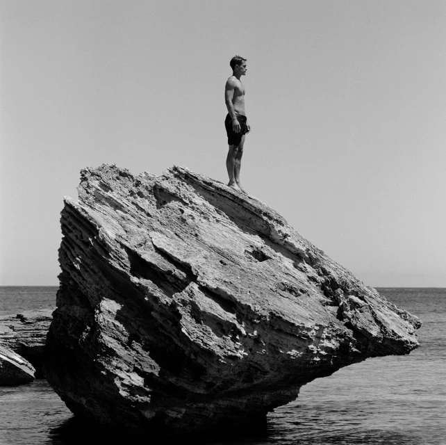 A shirtless man in black shorts stands atop a large rock formation in the ocean under a clear sky, a monochrome outdoor scene.