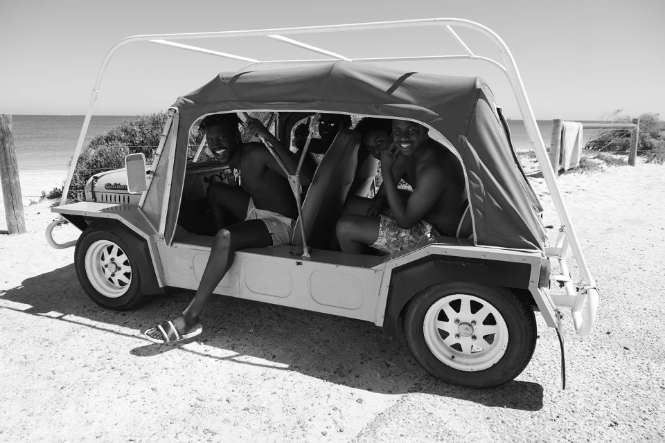 Black and white shot of three young adult men in a roofed, open-sided vehicle on a beach with ocean in the background.