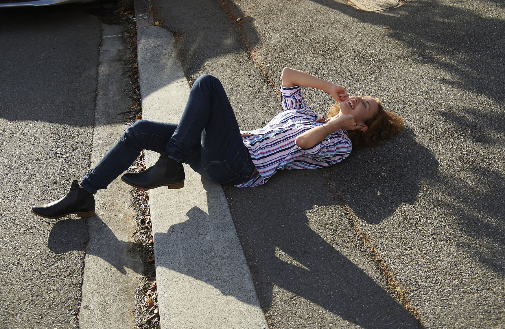 A young woman in jeans and a striped shirt lies on the asphalt next to a curb, smiling with her hands up near her face.