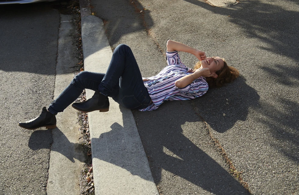 A young woman in jeans and a striped shirt lies on the asphalt next to a curb, smiling with her hands up near her face.