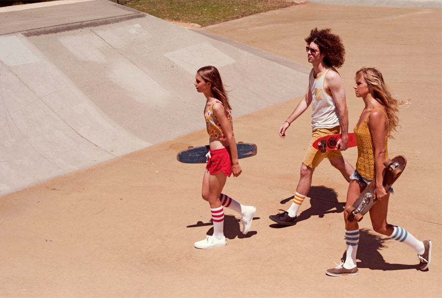 Three people walk across a tan concrete skatepark, carrying skateboards; the central figure has an afro and wears a tank top.