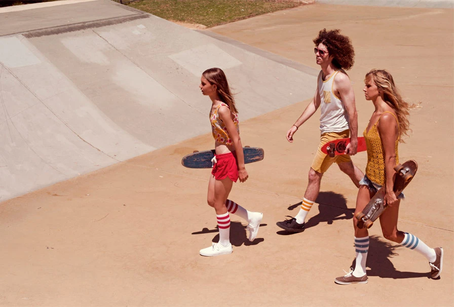 Three people walk across a tan concrete skatepark, carrying skateboards; the central figure has an afro and wears a tank top.
