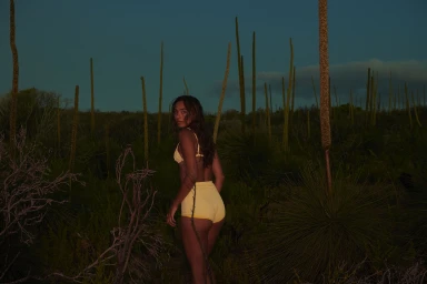 Woman in yellow two-piece swimwear standing amidst tall green flora under a twilight sky by Ian & Erick.