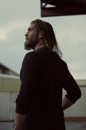 Man with long hair and a beard wearing a dark long-sleeve shirt outdoors against an overcast sky and building structures.