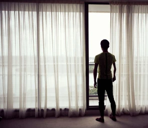 Man standing with his back to the camera looking out a large window framed by sheer white curtains.