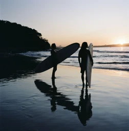 Two surfers standing on a wet sandy beach at sunset holding surfboards, with dark silhouettes against a glowing orange sky.