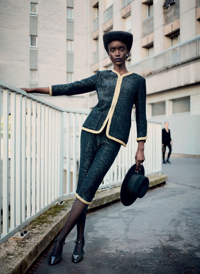 Dark-skinned model in a dark blue skirt suit with gold trim, holding a black hat, outdoors against an urban building backdrop.