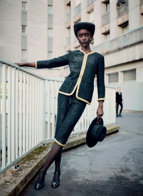 Dark-skinned model in a dark blue skirt suit with gold trim, holding a black hat, outdoors against an urban building backdrop.