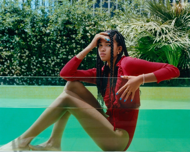 Young woman in red swimsuit and top sits in a swimming pool with greenery and sunlight creating a vibrant, summery fashion image.