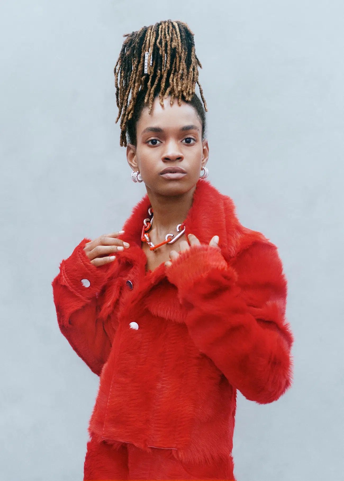 Portrait of a woman in a red faux fur jacket and necklace, with dreadlocks up in a high style against a solid grey background.