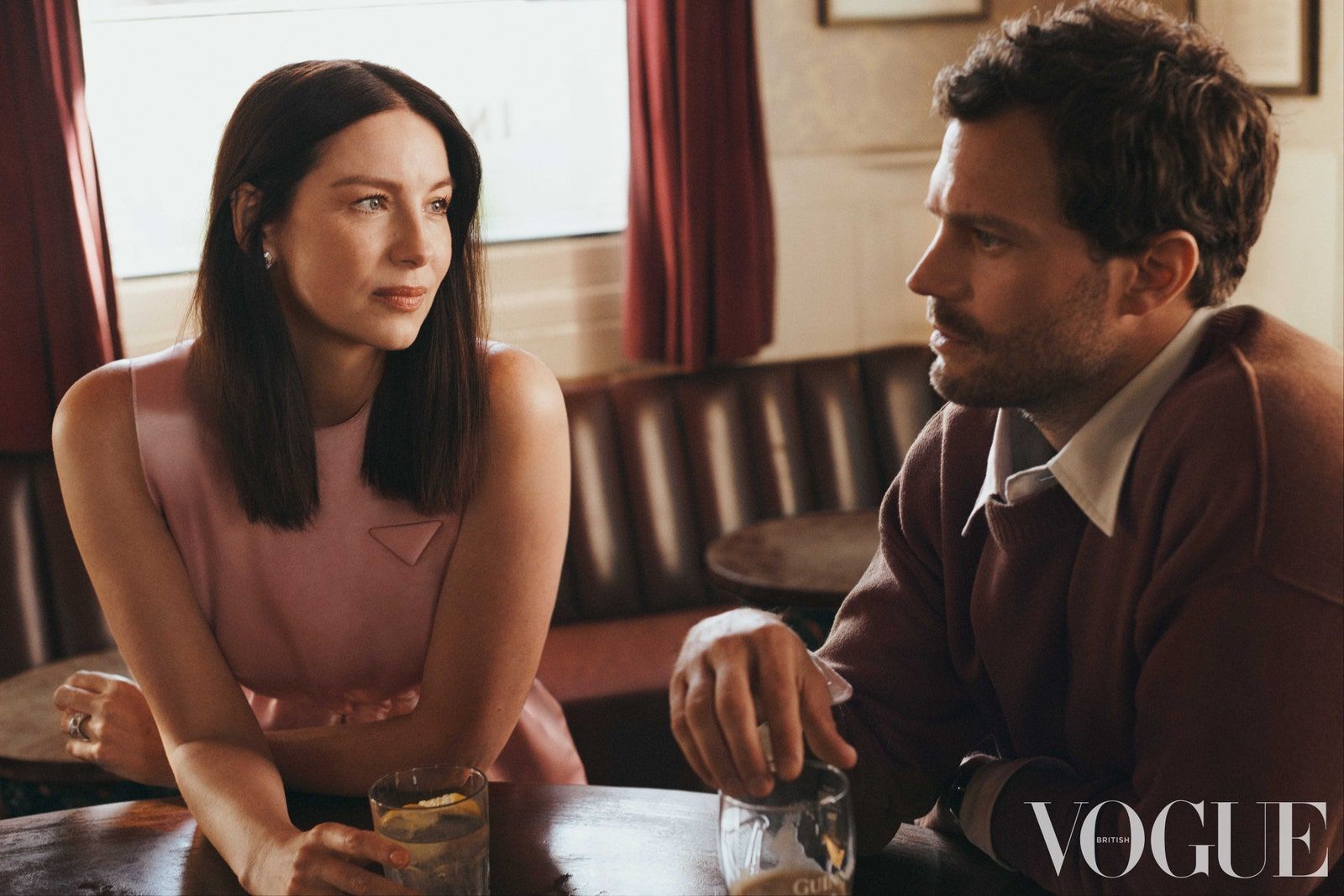 Caitriona Balfe and Jamie Dornan sit at a table in an indoor setting, with drinks, soft lighting, and a magazine in the foreground.