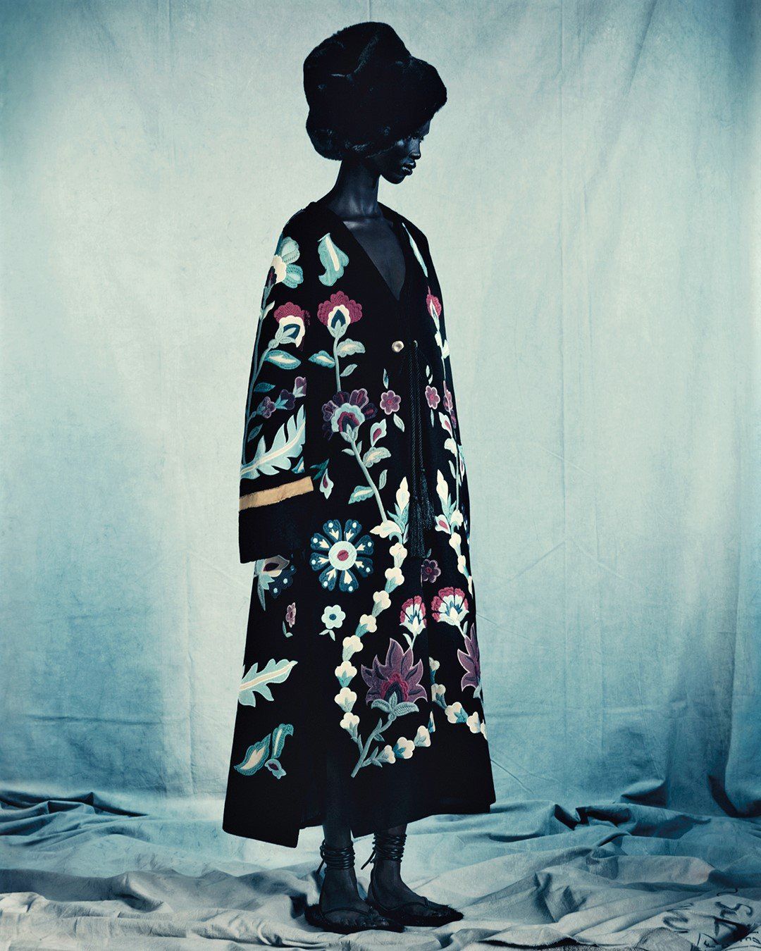 A fashion portrait of a Black woman wearing a dark floral coat and a fur hat against a textured backdrop in a studio setting.