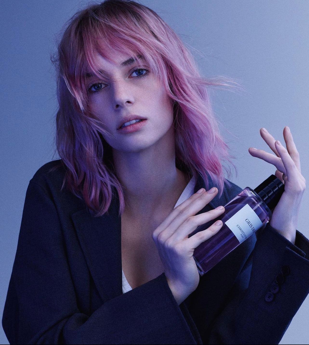Woman with pink hair poses indoors holding a glass perfume bottle, wearing a black blazer against a gradient blue background.