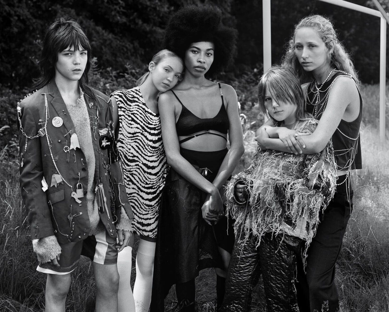 Group of five fashion models in a black and white outdoor shot, featuring diverse clothing styles and textures, with a background of grass and trees.