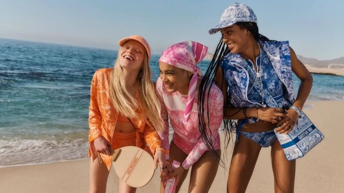Three smiling women in vibrant patterned outfits stand on a sandy beach next to turquoise ocean. Fashion and beachwear.