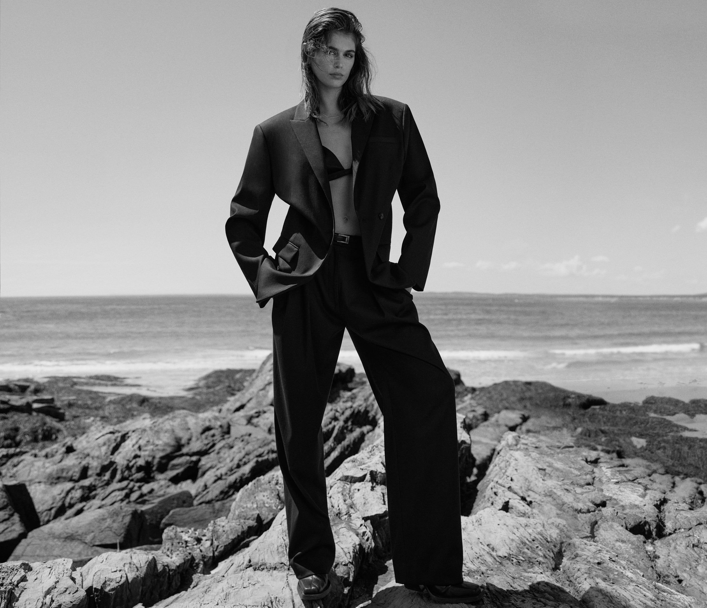 Monochrome fashion shot on rocky beach: adult woman posing in blazer and wide-leg pants with ocean backdrop under bright sky.