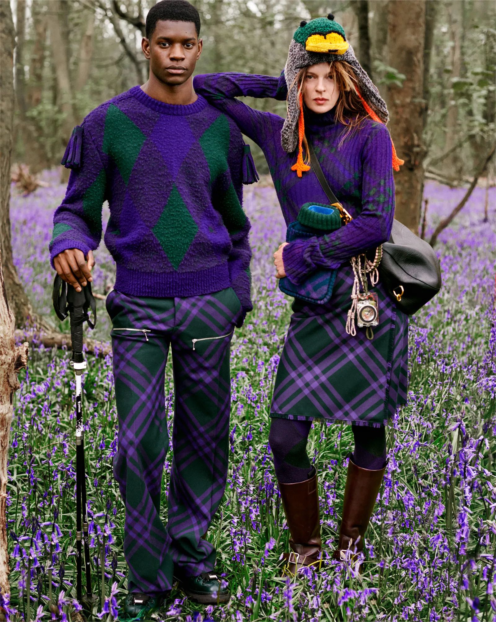 Two models pose outdoors in a forest clearing filled with purple flowers, wearing matching plaid and diamond-patterned sweaters.