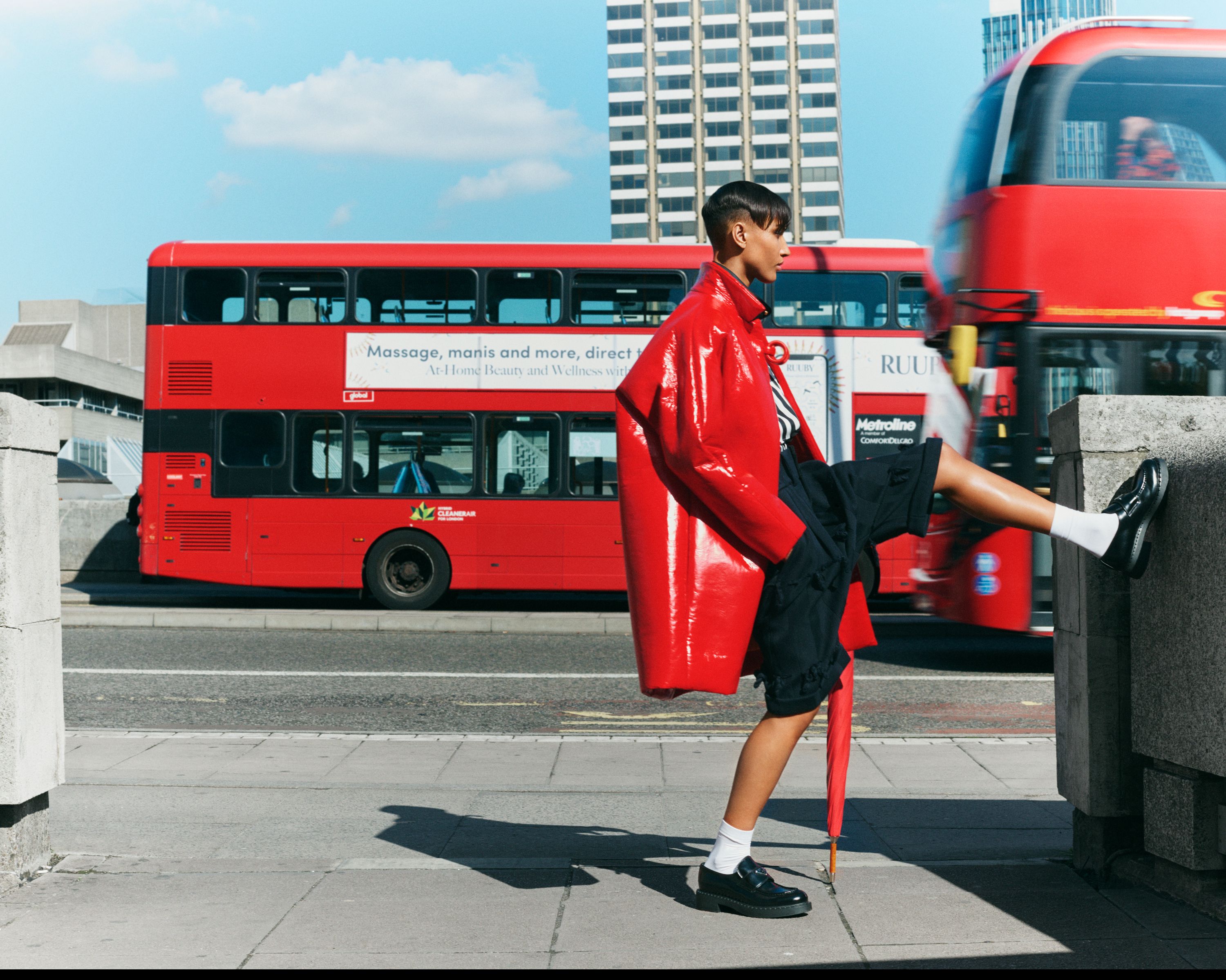 A woman in a red coat poses on a city street with a red double-decker bus blurred in the background on a sunny day.