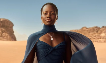 Lupita Nyongo wearing a navy blue dress and matching cape with silver diamond earrings and pendant necklace in a desert landscape
