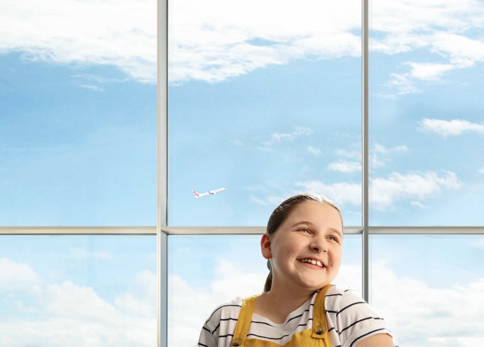 Young girl smiles while looking at an airplane through the airport window. Blue skies with clouds fill the background.