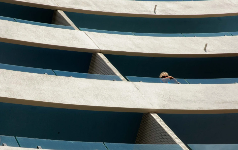 Woman stands on a curved apartment balcony in sunlight. Modern architecture features white concrete and blue railings in an urban setting.
