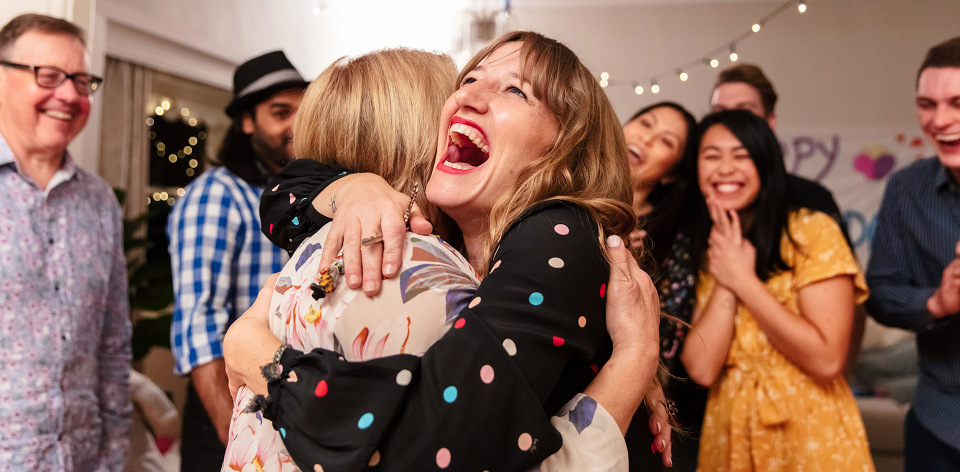 Two women embrace at an indoor party with string lights and diverse group of smiling and laughing people in the background.