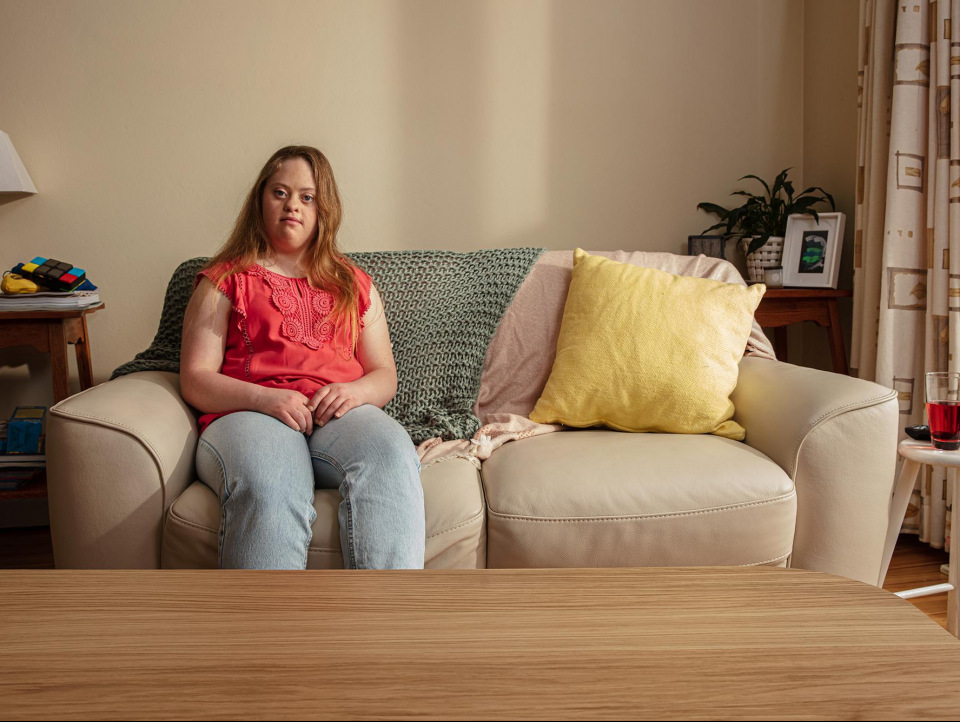 Woman with Down syndrome sits on a couch in a living room with a table in front, illuminated by natural indoor light.