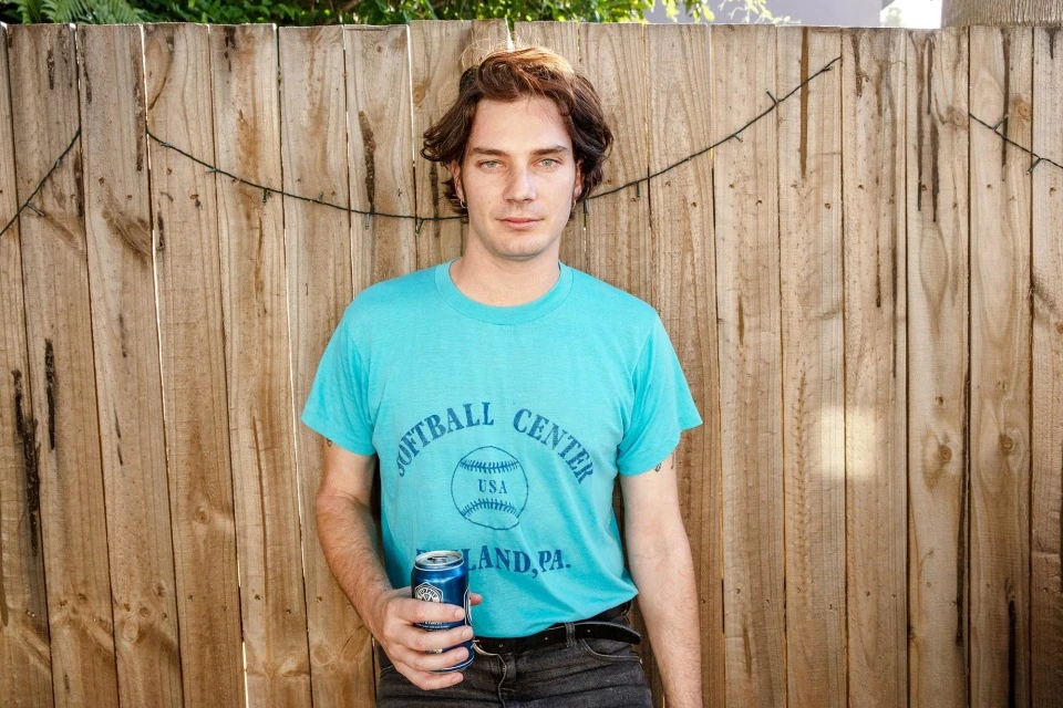 Young man outdoors stands in front of a wooden fence, wearing a vintage t-shirt, holding a blue beverage can, looking at the camera.