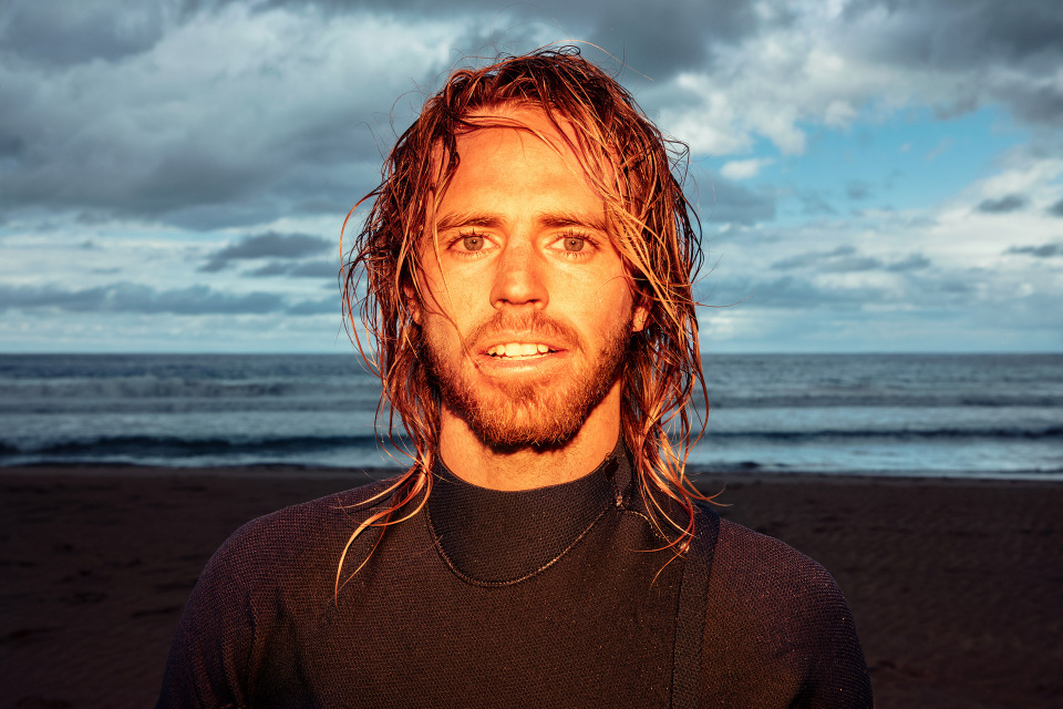 Portrait of a surfer with long blond hair and a wetsuit, smiling on a beach with ocean waves and cloudy sky in the background.