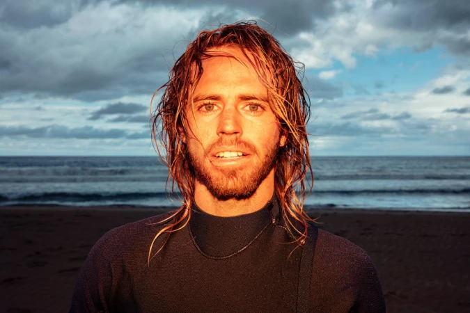 Portrait of a surfer with long blond hair and a wetsuit, smiling on a beach with ocean waves and cloudy sky in the background.