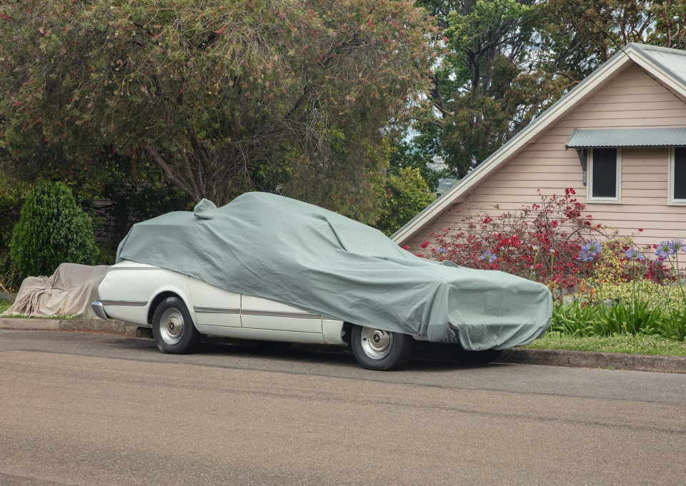 A white vintage car is covered with a green tarp on a residential street with a house and trees in the background.