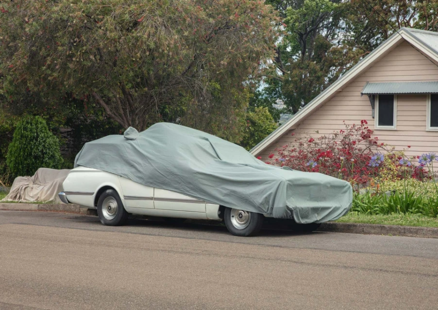 A white vintage car is covered with a green tarp on a residential street with a house and trees in the background.