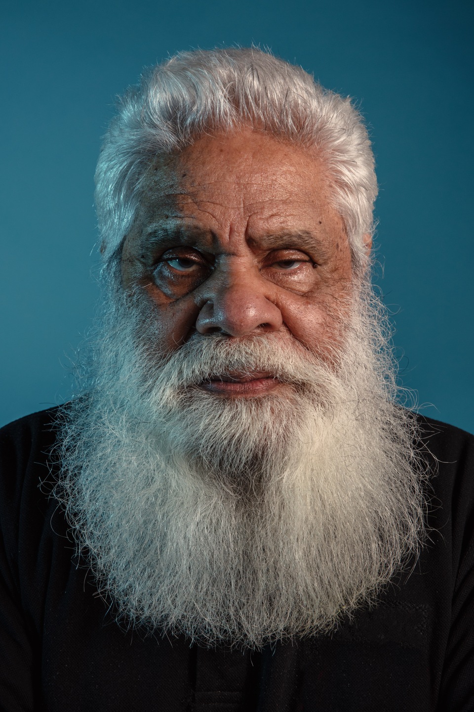 Portrait of an elderly man with a long white beard and gray hair, wearing a black shirt against a blue background in a studio setting.