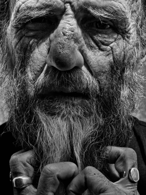 Striking black and white portrait of an elderly man with a long beard and weathered skin, his hands visible with rings.