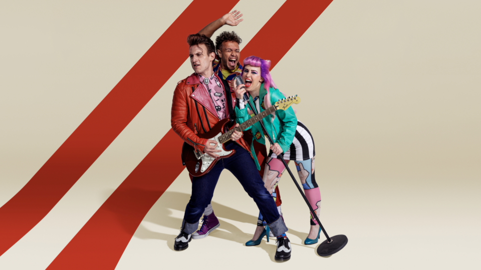Music band performs with a guitarist in a red jacket, a vocalist in a turquoise jacket, and a background vocalist with hand raised.