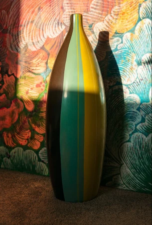 Tall striped ceramic vase standing against a brightly patterned floral wallpaper backdrop with shadow
