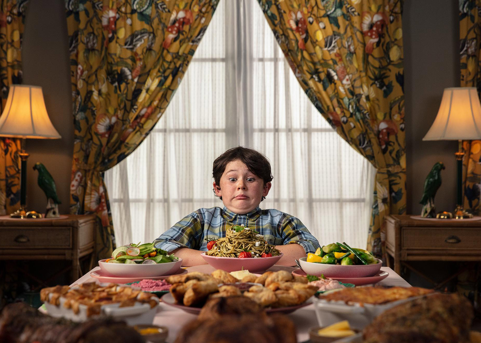 Boy with an overwhelmed expression sits at a table laden with various food dishes, featuring salads, pasta, and pastries in an indoor setting.