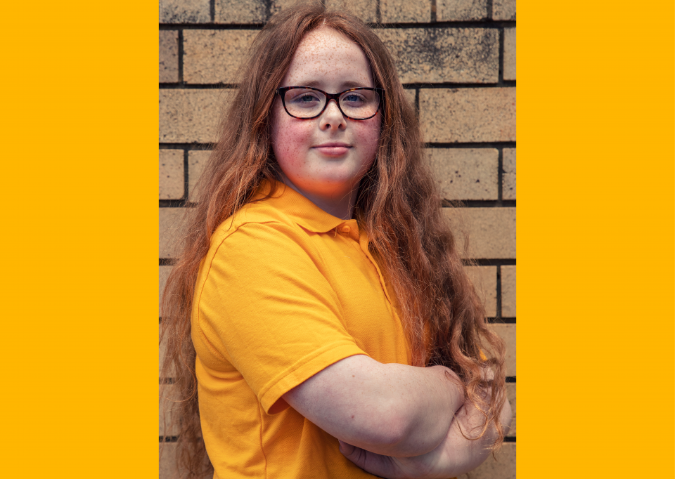 A young woman with long red hair and glasses stands with her arms crossed in front of a brick wall, wearing a yellow polo shirt.