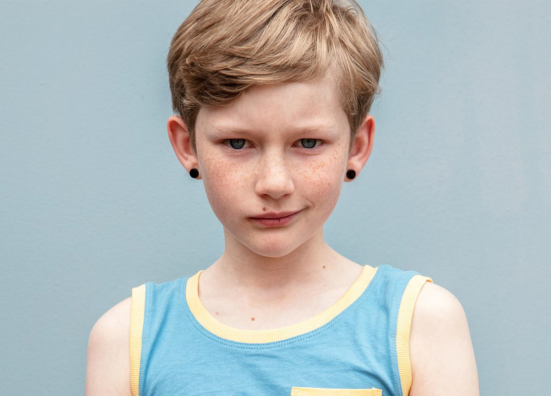 Close-up studio shot of a freckled child with blonde hair wearing a blue tank top against a light blue background.
